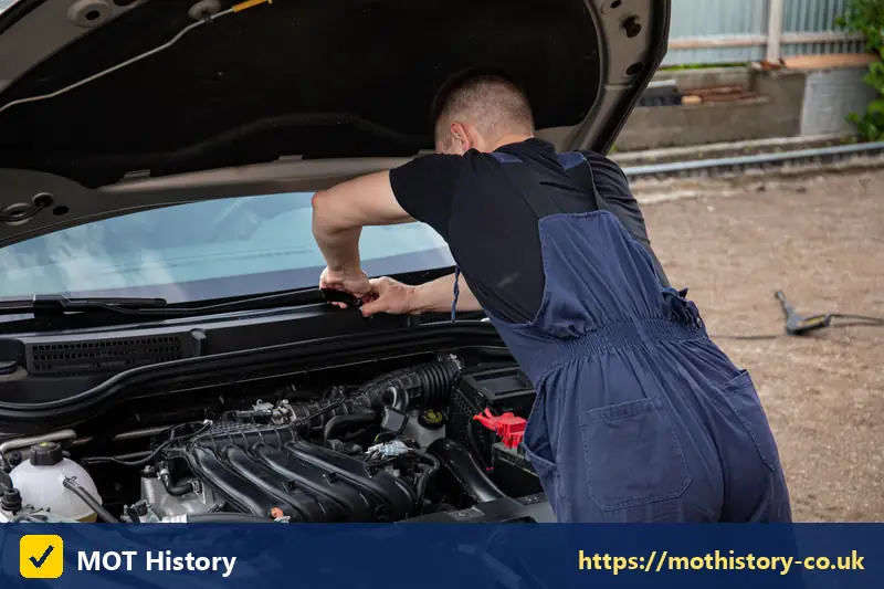 Mechanic in blue overalls leaning over an open bonnet - what does mot test